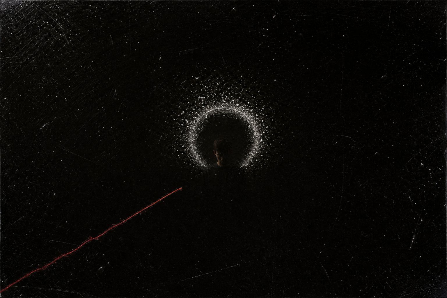 A distant head surrounded by a ring of pale dust and static on black glass while a red thread stops short.
